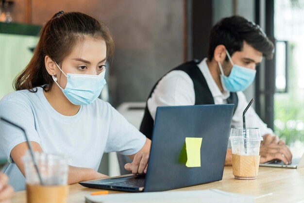 Il nuovo concetto di stile di vita normale, gli uomini d'affari che indossano la maschera si siedono con l'allontanamento sociale lavorando in aree pubbliche o in uno spazio di lavoro condiviso con laptop e smartphone