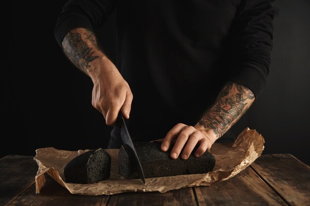 Il panettiere irriconoscibile con le mani tatuate ha tagliato il pane al carbone di legna fatto in casa appena sfornato con il grande coltello principale sulle fette sulla carta del mestiere sul tavolo rustico in legno