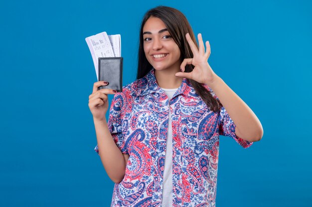Il passaporto turistico della tenuta della giovane bella donna con i biglietti che sorridono allegramente che fa bene firma sopra la parete blu isolata