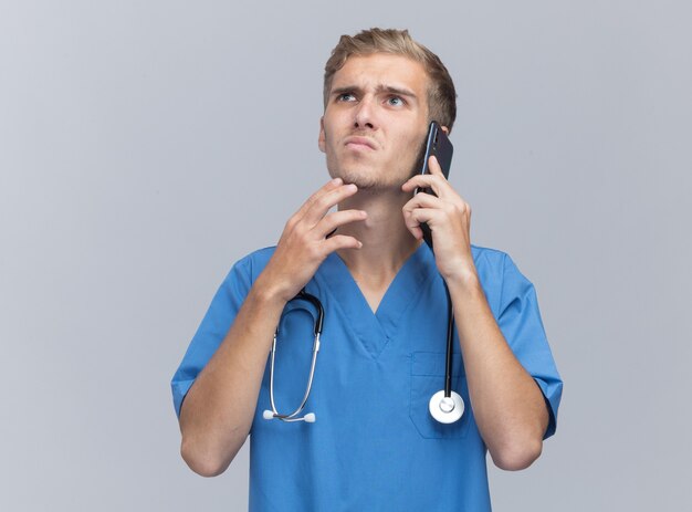 Il pensiero che osserva in su il giovane medico maschio che porta l'uniforme del medico con lo stetoscopio parla sul telefono che mette la mano sul mento isolato sulla parete bianca