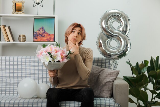 Il pensiero ha afferrato il mento bel ragazzo durante la felice giornata delle donne con in mano un bouquet seduto sul divano in soggiorno