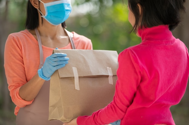 Il postino, il fattorino che indossa la maschera porta una piccola scatola consegnata al cliente davanti alla porta di casa. la donna che indossa la maschera previene il covid 19, l'epidemia di infezione da coronavirus. concetto di acquisto di consegna a domicilio.