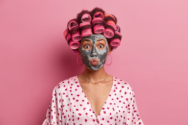 Il primo piano del volto di bella giovane donna mantiene le labbra arrotondate, applica una maschera di bellezza, indossa i bigodini, vestita con una veste di seta casual, isolata sopra il rosa. Routine di bellezza e concetto di acconciatura