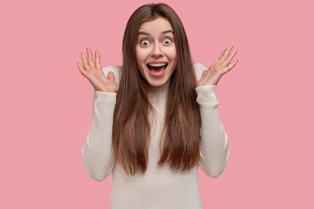 Il primo piano del volto di una donna eccitata attraente allarga le palme vicino al viso, ha un'espressione sorpresa, ha i capelli lunghi, vestito con indifferenza