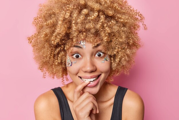 Il primo piano del volto di una giovane donna felice e sorpresa che morde il dito sembra chiesta abbia il viso decorato con pietre lucenti ha capelli ricci naturali vestiti con una maglietta nera casual isolata su sfondo rosa