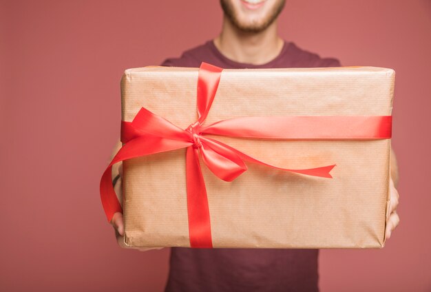 Il primo piano dell&#39;uomo che tiene il contenitore di regalo avvolto carta marrone con l&#39;arco rosso del nastro