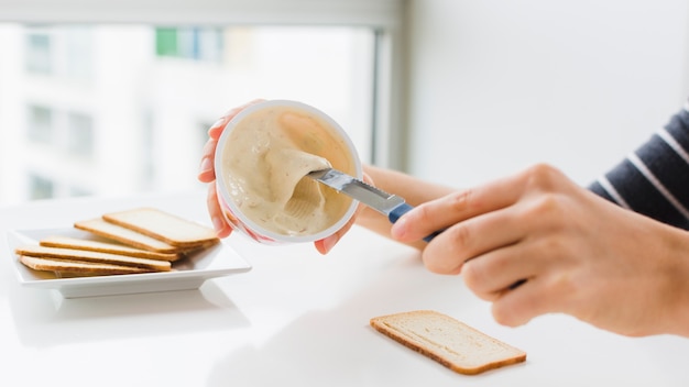 Il primo piano della donna che applica il formaggio si è sparso su pane con il coltello sopra la tavola bianca