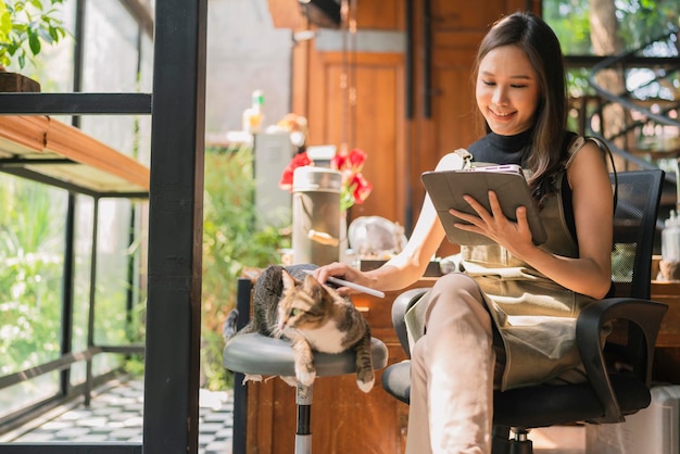 Il progettista creativo della femmina adulta asiatica casual si rilassa lavorando con il suo fine settimana mattutino dell'animale domestico del gatto a casa studioyoung asiatico asiatico indossa il grembiule ricerca le informazioni per il suo laboratorio tramite il dispositivo tablet