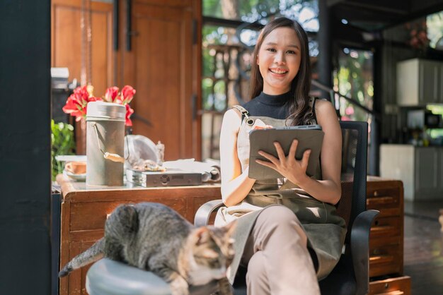 Il progettista creativo della femmina adulta asiatica casual si rilassa lavorando con il suo fine settimana mattutino dell'animale domestico del gatto a casa studioyoung asiatico asiatico indossa il grembiule ricerca le informazioni per il suo laboratorio tramite il dispositivo tablet