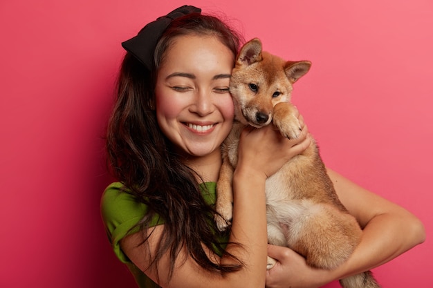 Il proprietario dell'animale domestico della donna felice tiene il cane sveglio vicino al fronte, trasporta il cucciolo di shiba inu, sorride piacevolmente, gode del momento dolce, isolato sopra fondo rosa.