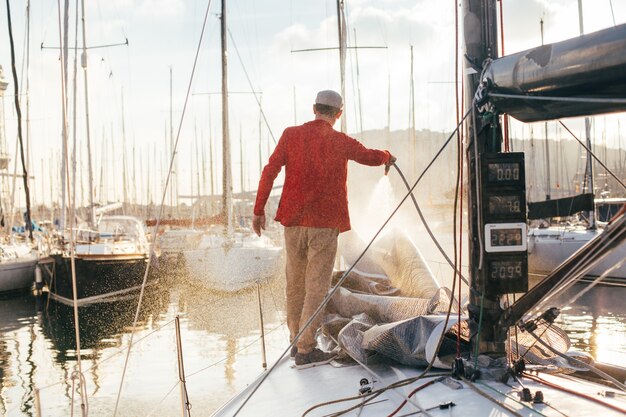 Il proprietario della barca a vela o il diportista utilizza il tubo flessibile per lavare l'acqua salata dal ponte dello yachr quando è attraccato o parcheggiato nel porto turistico al tramonto