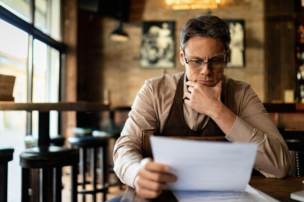 Il proprietario di un caffè pensieroso legge mentre esamina le scartoffie e legge i rapporti dopo l'orario di lavoro.