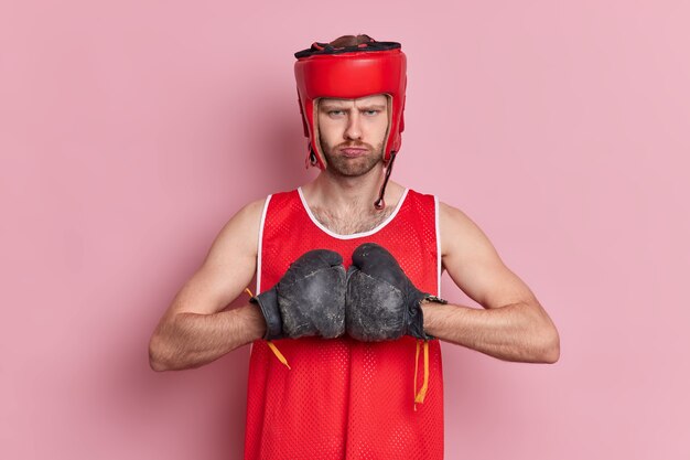 Il pugile serio e forte con la barba lunga indossa cappello e guantoni da boxe tiene le mani unite si prepara per la competizione sportiva pronta a combattere.