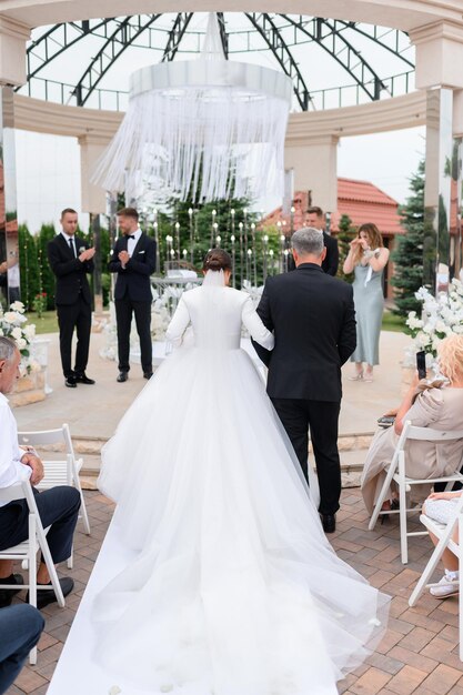 Il punto di vista posteriore del padre amorevole con la figlia della sposa in abito bianco lungo e gonfio va al goom durante la cerimonia di matrimonio all'aperto Momento toccante per gli ospiti e la coppia sposata Elegante altare nuziale