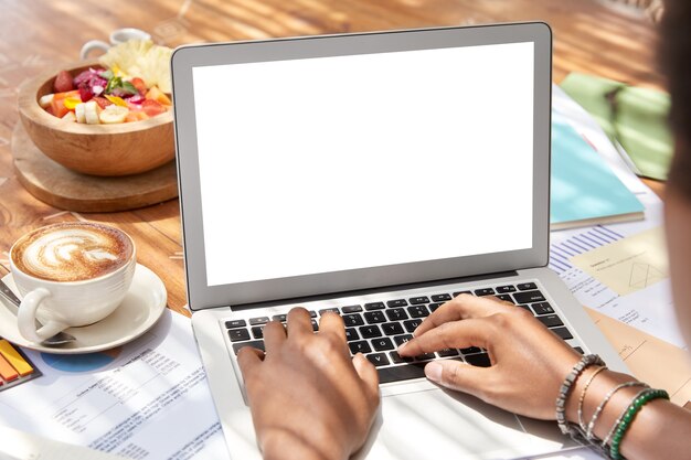 Il punto di vista posteriore della donna dalla pelle scura si siede davanti al computer portatile aperto con lo schermo bianco vuoto in bianco per la vostra pubblicità