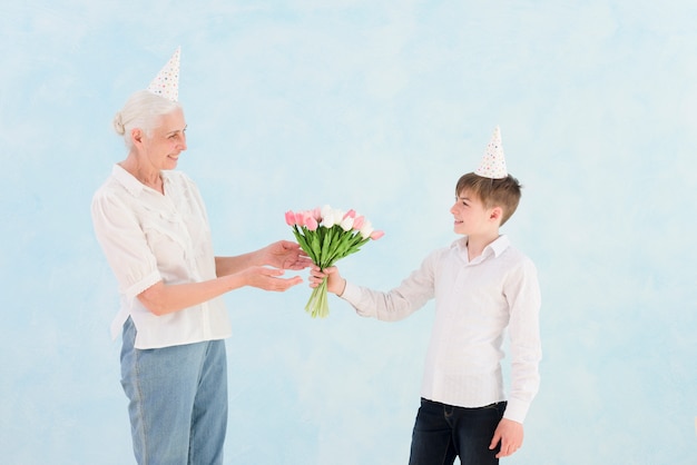 Il ragazzino felice che dà il tulipano fiorisce il mazzo a sua nonna contro il contesto blu