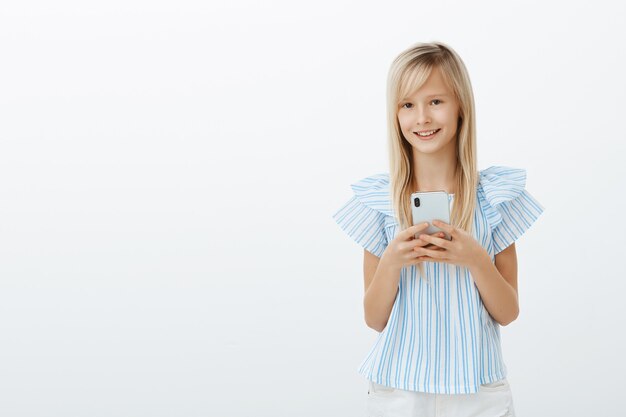 Il ragazzino ha rubato il cellulare di papà per giocare. Ritratto di affascinante ragazza giovane felice con capelli biondi, tenendo lo smartphone e sorridente ampiamente, guardando cartoni animati o messaggistica con gli amici oltre il muro grigio