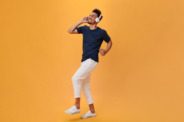 Il ragazzo alla moda si muove ritmicamente e si diverte con la musica in cuffia Uomo in pantaloni bianchi e camicia blu che balla su sfondo arancione