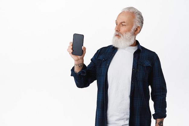 Il ragazzo anziano sembra sorpreso dallo schermo dello smartphone, mostrando l'applicazione del telefono cellulare o la pagina web sul display, in piedi stupito contro il muro bianco