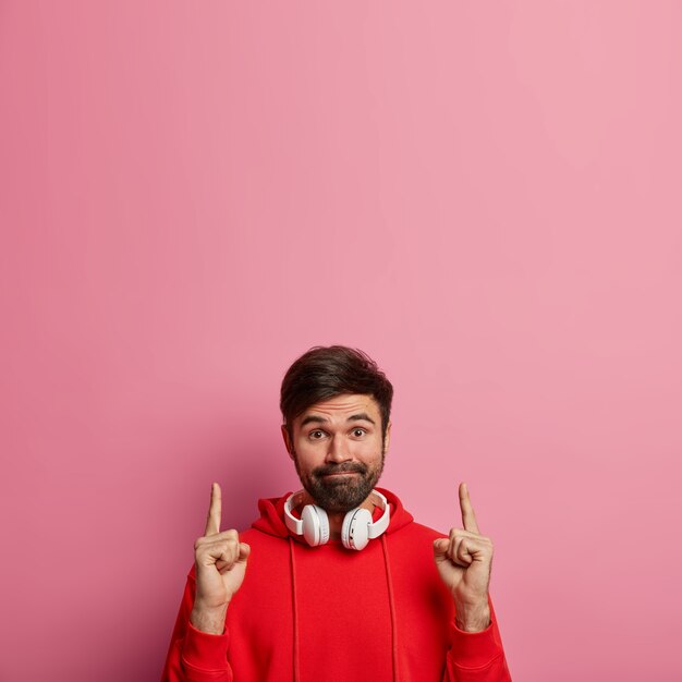 Il ragazzo barbuto hipster punta entrambi gli indici sopra, mostra uno spazio vuoto incredibile, fa una bella offerta, preme le labbra, indossa cuffie stereo e una felpa con cappuccio rossa isolata sul muro rosa pastello. Guarda in alto