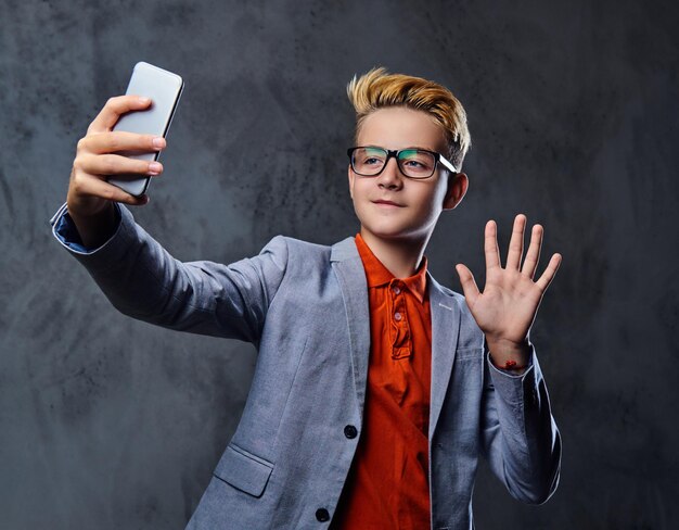 Il ragazzo biondo dell'adolescente in una giacca e occhiali da vista tiene lo smartphone.