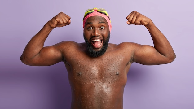 Il ragazzo dalla pelle scura felice e divertente mostra i muscoli dopo il nuoto, dimostra un corpo forte e nudo bagnato, ha una folta barba