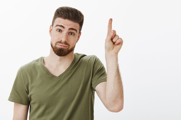 Il ragazzo ha un suggerimento di alzare il dito indice per aggiungere un'idea in piedi sciocco e carino con un'espressione esitante e timida che fa il broncio inclinando la testa e guardando con un sorriso carino, rivolto verso l'alto sul muro bianco