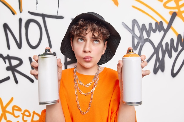 Il ragazzo hipster premuroso concentrato sopra tiene due bottiglie di aerosol indossa un cappello nero e una maglietta arancione disegna un muro di graffiti in un luogo pubblico ama l'arte di strada