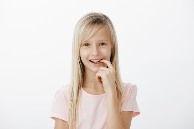 Il ragazzo intelligente creativo ha inventato una grande idea. Ritratto di ragazza carina sognante con capelli biondi, sorridente con gioia e dito mordace, che ha un piano o vuole fare qualcosa di intrigante, in piedi sopra il muro grigio