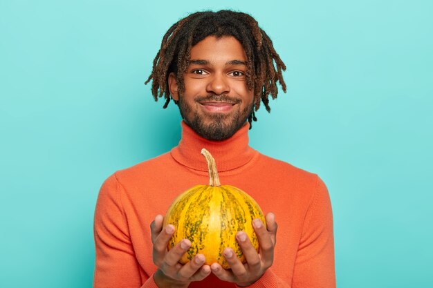 Il ragazzo soddisfatto con i dreadlocks tiene una piccola zucca, sta per scolpire la festa di Halloween, ha barba e baffi, indossa un comodo poloneck