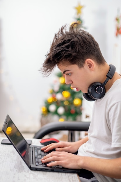 Il ragazzo teenager sta usando il computer portatile con le cuffie a casa. Albero di Natale sul muro. Faccia stupita