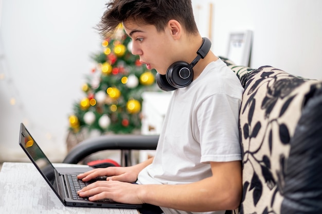 Il ragazzo teenager sta usando il computer portatile con le cuffie a casa. Albero di Natale sul muro. Faccia stupita