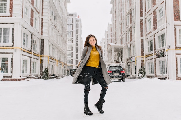 Il ritratto a figura intera della donna graziosa indossa un maglione giallo alla moda che scherza sulla strada sotto la nevicata. Foto all'aperto di donna europea sottile in cappotto caldo che balla sulla strada urbana.