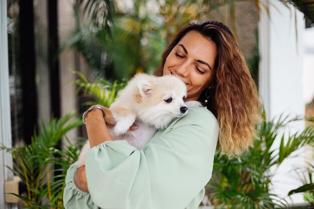 Il ritratto all'aperto della donna abbronzata europea riccia tiene lo spitz pomeranian del cane da compagnia felice