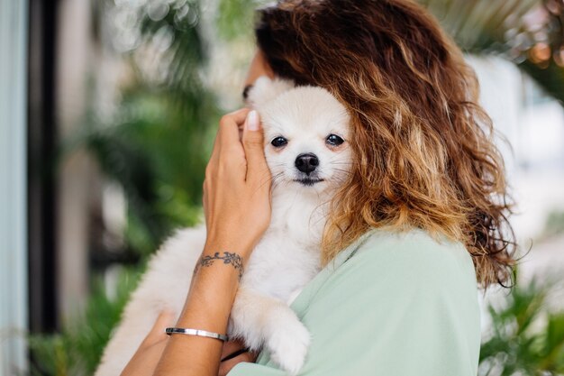 Il ritratto all'aperto della donna abbronzata europea riccia tiene lo spitz pomeranian del cane da compagnia felice