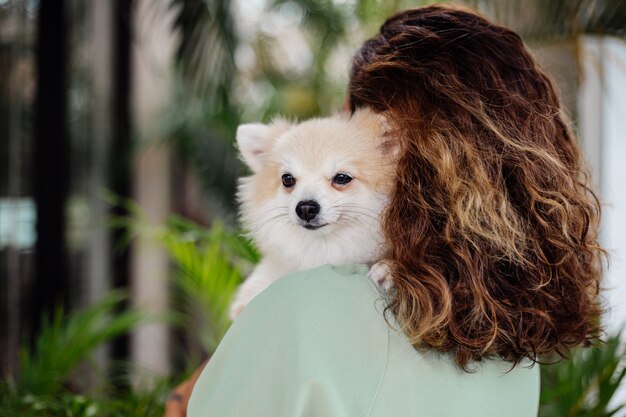 Il ritratto all'aperto della donna abbronzata europea riccia tiene lo spitz pomeranian del cane da compagnia felice