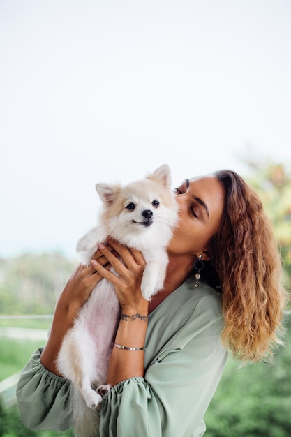 Il ritratto all'aperto della donna abbronzata europea riccia tiene lo spitz pomeranian del cane da compagnia felice