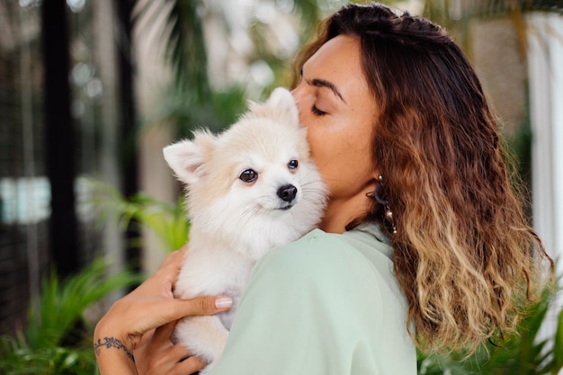 Il ritratto all'aperto della donna abbronzata europea riccia tiene lo spitz pomeranian del cane da compagnia felice