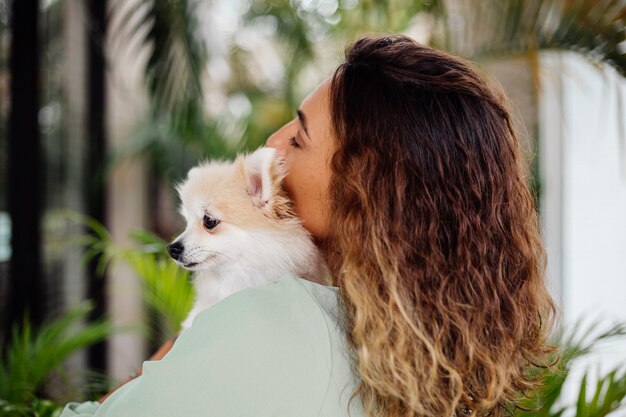 Il ritratto all'aperto della donna abbronzata europea riccia tiene lo spitz pomeranian del cane da compagnia felice