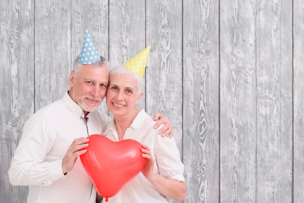 Il ritratto del cappello d&#39;uso del compleanno delle coppie senior felici che tiene il rosso sente il pallone di forma nel fondo di legno anteriore