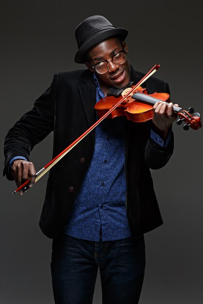 Il ritratto del giovane uomo sorridente nero bello in cappello che gioca il violino sull'oscurità