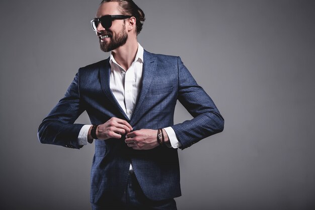 il ritratto del modello dell'uomo d'affari alla moda dei pantaloni a vita bassa alla moda bello si è vestito in vestito blu elegante in occhiali da sole che posano sul gray