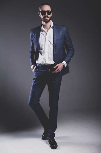 il ritratto del modello dell'uomo d'affari alla moda dei pantaloni a vita bassa alla moda bello si è vestito in vestito blu elegante in occhiali da sole che posano sul gray