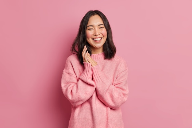 Il ritratto del modello femminile asiatico brunetta sincero strofina le palme e sorride ampiamente esprime la felicità si sente gioioso ha denti bianchi anche indossa un maglione casual oversize