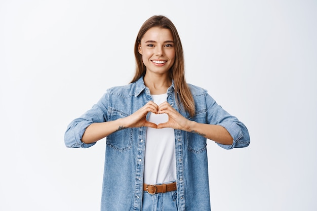 Il ritratto del modello femminile con i capelli biondi e il sorriso bianco ama qualcosa, mostrando il gesto del cuore e sorridendo per esprimere simpatia, parete dello studio