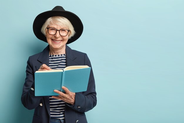 Il ritratto del pensionato femmina soddisfatto scrive la strategia del piano nel diario, ha un bell'aspetto intelligente, indossa occhiali e cappello nero, isolato sopra la parete blu con spazio vuoto. Imprenditrice con blocco note