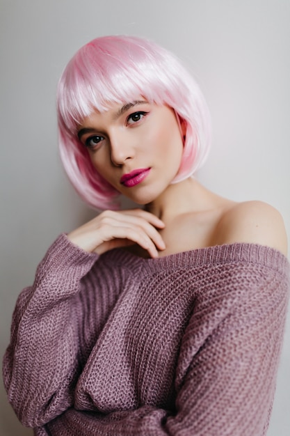 Il ritratto del primo piano del modello femminile caucasico adorabile indossa il peruke colorato elegante. Splendida signora con capelli rosa chiaro in posa sognante su una parete chiara.