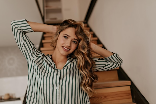 Il ritratto del primo piano della donna riccia bionda abbastanza giovane tocca i suoi lunghi capelli ondulati esamina la macchina fotografica