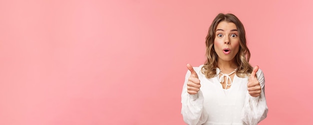 Il ritratto del primo piano della ragazza bionda impressionata che mostra il thumbsup sembra stupito e incredulo come processin