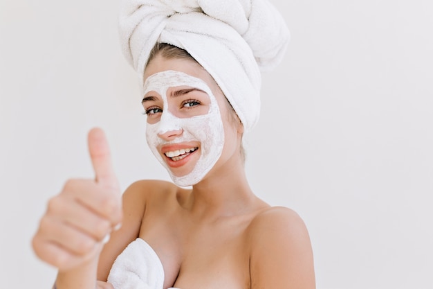 Il ritratto del primo piano di bella giovane donna che sorride con gli asciugamani dopo fare il bagno fa la maschera cosmetica sul suo viso.